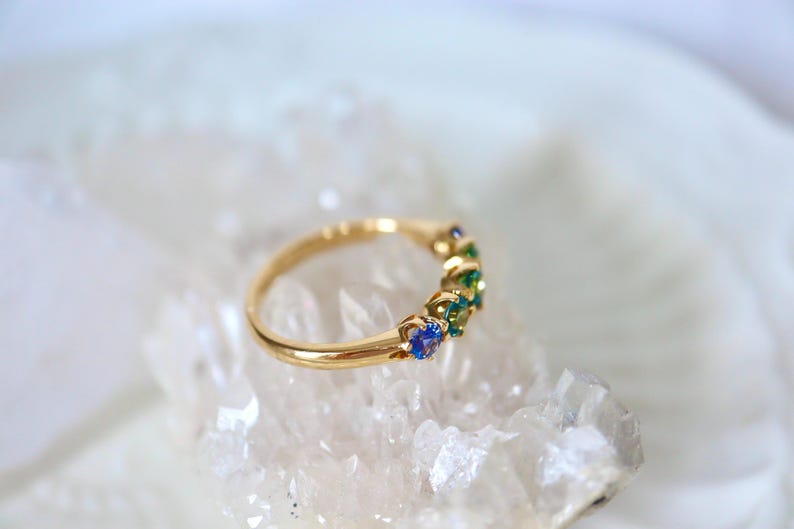 May include: A gold band ring with a row of blue and green gemstones. The ring is set on a white crystal surface.