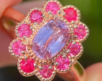 May include: A gold ring with a central, pale purple, oval gemstone, encircled by a halo of smaller, bright pink gemstones. The ring has a floral design.