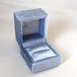 May include: A small, open, blue velvet ring box. The box is rectangular with a hinged lid and a plush interior designed to hold a ring. The box is a soft, muted blue color.