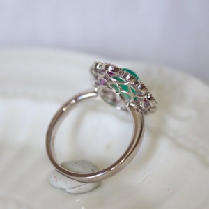 May include: A silver ring with a green gemstone surrounded by a delicate, intricate design of smaller gemstones. The ring is set on a white surface.