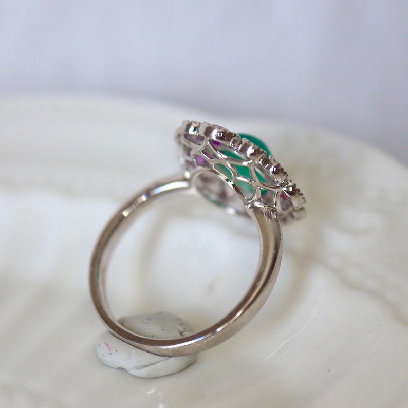May include: A silver ring with a green gemstone surrounded by a delicate, intricate design of smaller gemstones. The ring is set on a white surface.