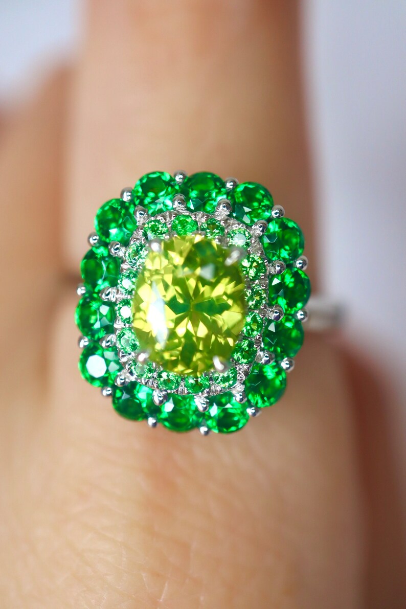 May include: A ring with a large, yellow-green gemstone surrounded by a double halo of smaller green gemstones.