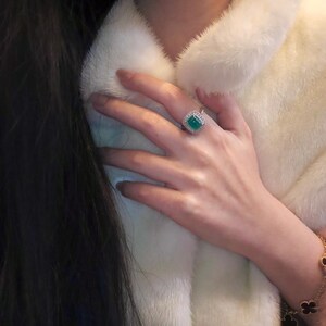 May include: A white fur jacket with a green emerald ring on a finger. The ring has a square emerald stone surrounded by smaller diamonds.