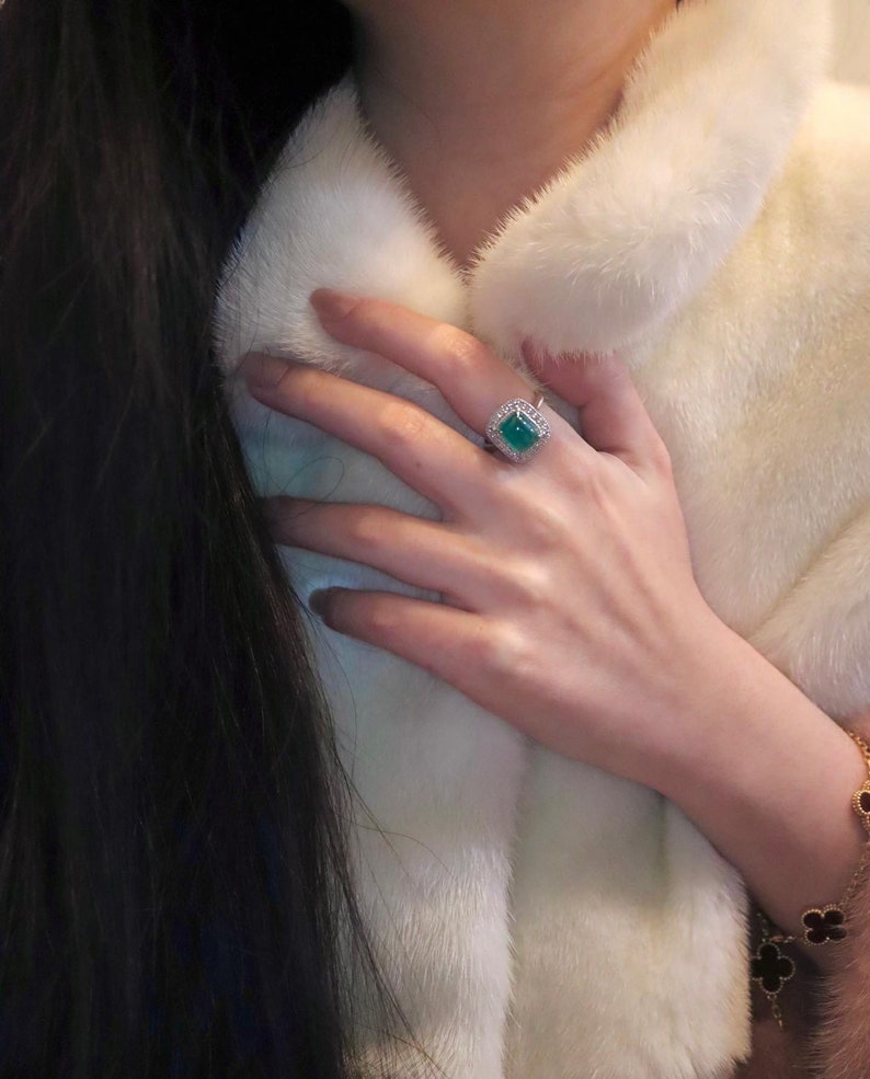 May include: A white fur jacket with a green emerald ring on a finger. The ring has a square emerald stone surrounded by smaller diamonds.