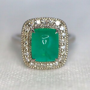 May include: A silver ring with a large, square, green gemstone surrounded by a halo of smaller, white gemstones.