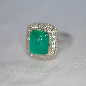 May include: A silver ring with a square emerald green gemstone surrounded by a halo of smaller clear gemstones.