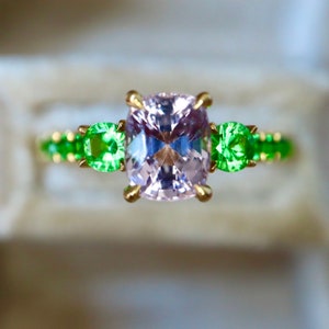 May include: A gold ring with a large, light pink, cushion-cut gemstone surrounded by smaller, green gemstones.