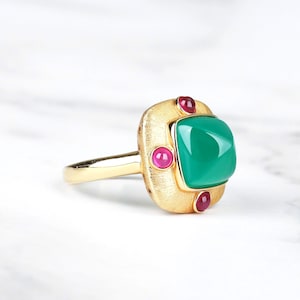 May include: A gold ring featuring a square green gemstone surrounded by four smaller red gemstones.