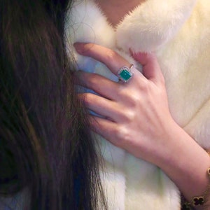 May include: A woman's hand wearing a ring with a large, square, emerald green gemstone surrounded by smaller diamonds. The ring is set in silver.