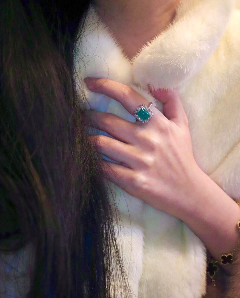 May include: A woman's hand wearing a ring with a large, square, emerald green gemstone surrounded by smaller diamonds. The ring is set in silver.