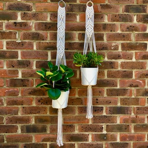 May include: Two white macrame plant hangers with wooden rings hanging from a brick wall. Each hanger holds a white pot with a green plant.