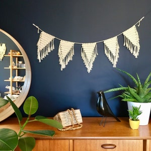 Puede incluir: Un banderín de macramé blanco con formas triangulares cuelga de una pared azul oscuro. Una figurita de pájaro negro se encuentra sobre una mesa de madera con un libro tejido y una planta en maceta.