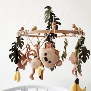 May include: A baby mobile featuring felt monkeys, bananas, and green leaves suspended from a wooden hoop. The mobile has a jungle theme, with the monkeys in various poses. The bananas are yellow and the leaves are dark green.
