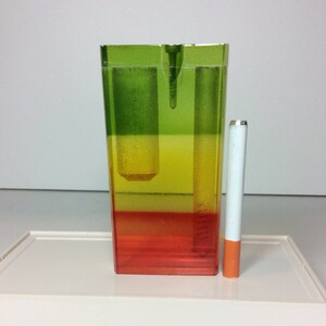 May include: A rectangular ashtray with a layered color design of green, yellow, and red. A cigarette with a white filter and orange tip stands next to the ashtray on a white tray.