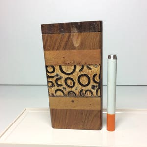 May include: A wooden box with a geometric design and a cigarette. The box is made of various wood tones and features a section with black circles. The cigarette has a white filter and an orange tip.