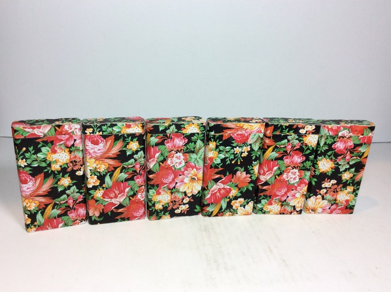 Puede incluir: Seis peque&ntilde;as cajas negras con un patr&oacute;n floral en tonos rojos, naranjas, amarillos y verdes. Las cajas est&aacute;n alineadas en una fila.