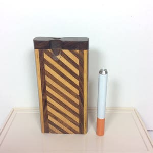 May include: A wooden box with a striped pattern and a small hole in the top. The box is about 10 cm tall and 5 cm wide. It is sitting on a white surface next to a silver and orange metal tube.