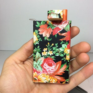 Puede incluir: Una peque&ntilde;a caja negra con estampado floral, una pipa de metal y una tapa de madera. La caja est&aacute; siendo sostenida por una mano.