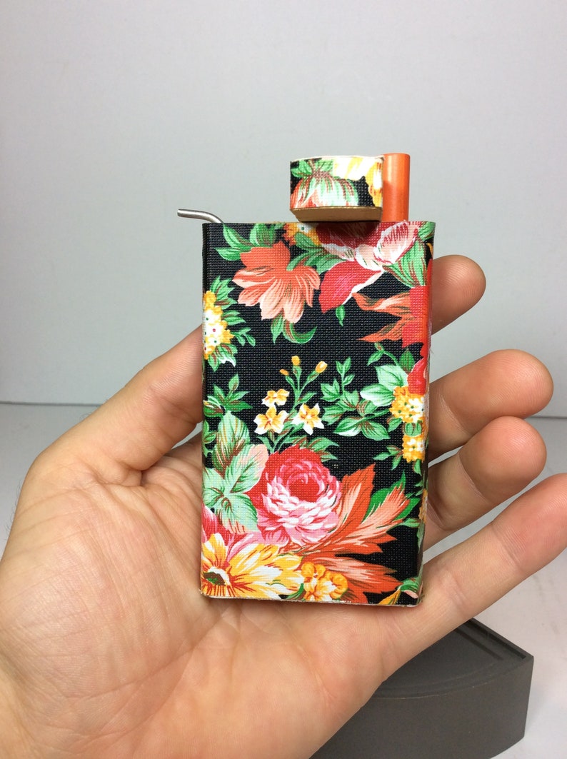 Puede incluir: Una peque&ntilde;a caja negra con estampado floral, una pipa de metal y una tapa de madera. La caja est&aacute; siendo sostenida por una mano.