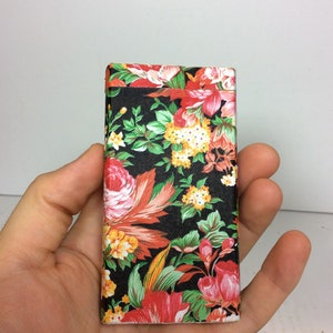Puede incluir: Una peque&ntilde;a caja rectangular cubierta de una tela negra con un patr&oacute;n floral. El patr&oacute;n presenta flores rojas, rosas, amarillas y verdes sobre un fondo negro.