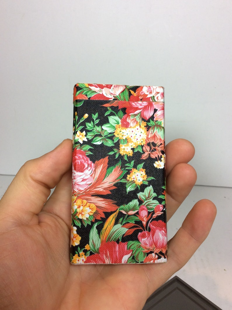 Puede incluir: Una peque&ntilde;a caja rectangular cubierta de una tela negra con un patr&oacute;n floral. El patr&oacute;n presenta flores rojas, rosas, amarillas y verdes sobre un fondo negro.