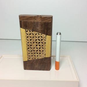 May include: A wooden box with a geometric design and a cigarette. The box is made of dark brown and tan wood with a decorative floral pattern. The cigarette has a white filter and an orange tip.