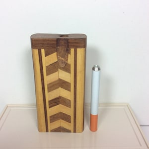 May include: A wooden box with a chevron pattern on the front. The box is closed and has a small hole on the top. A cigarette is next to the box for size comparison.
