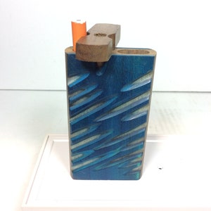 May include: A blue wooden box with a carved design and a wooden lid. The lid has a small hole for a cigarette. The box is sitting on a white surface.