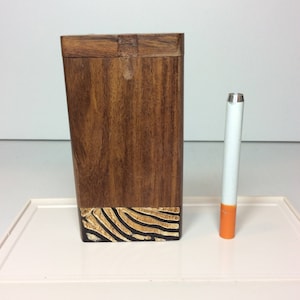 May include: A wooden cigarette case with a zebra stripe design at the base. The case is brown and rectangular, with a hinged lid. A single cigarette with a white filter and orange tip is next to the case.