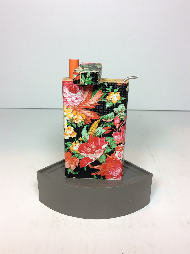 Puede incluir: Una caja de madera negra con estampado floral, tapa y pipa de metal. La caja est&aacute; sobre una base gris curva.