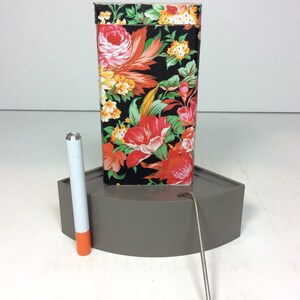 Puede incluir: Una caja de hojalata con estampado floral con una varilla de metal plateado y un tubo blanco y naranja. La caja de hojalata est&aacute; sobre una base gris.
