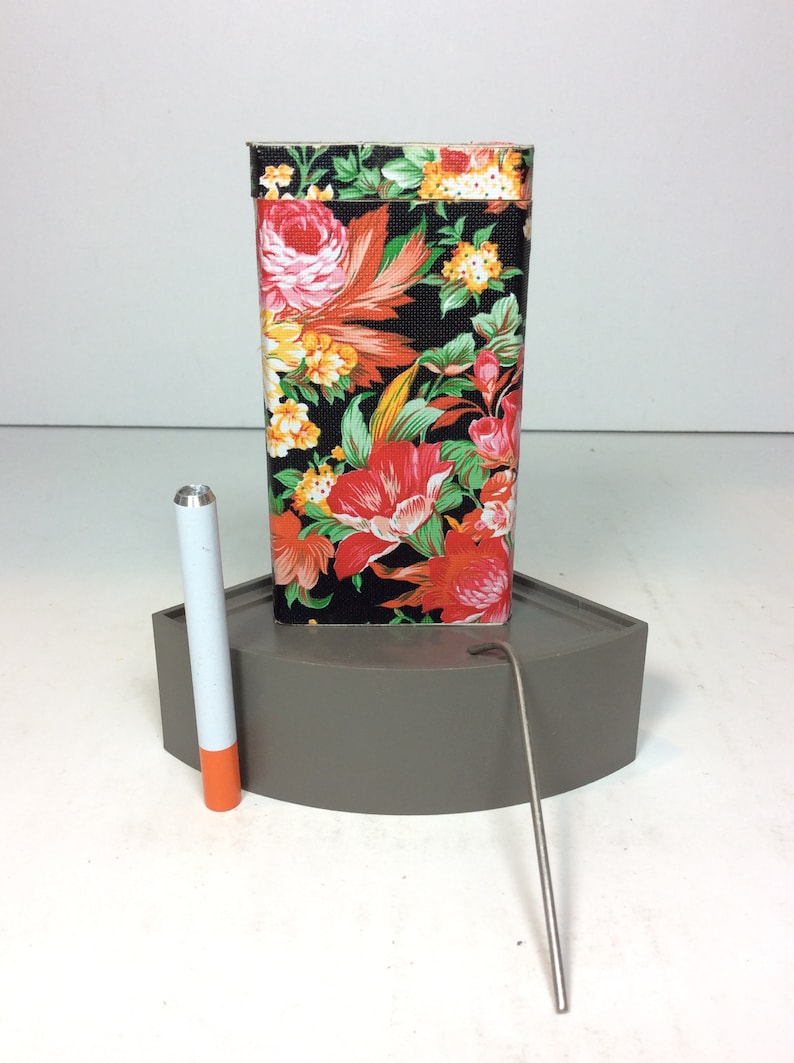 Puede incluir: Una caja de hojalata con estampado floral con una varilla de metal plateado y un tubo blanco y naranja. La caja de hojalata est&aacute; sobre una base gris.