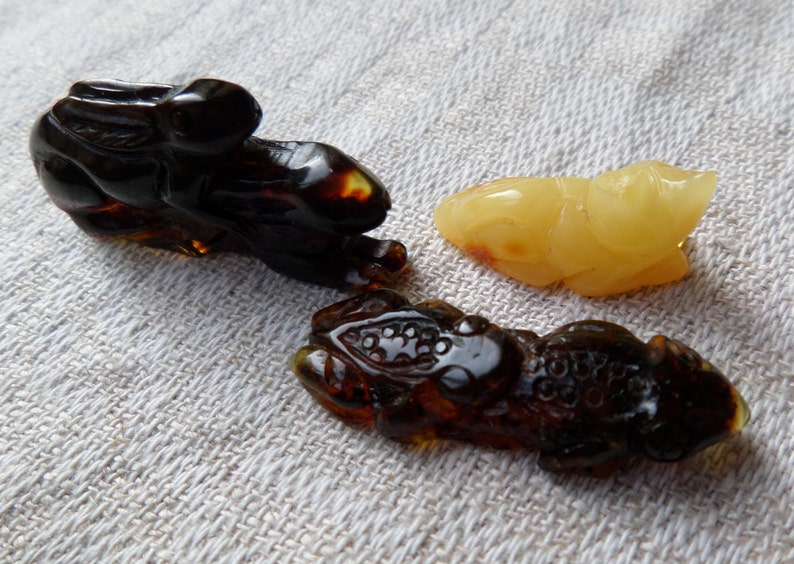 May include: Three amber carvings of animals: a rabbit, a frog, and a fish. The rabbit and frog are dark amber, while the fish is a lighter yellow amber.