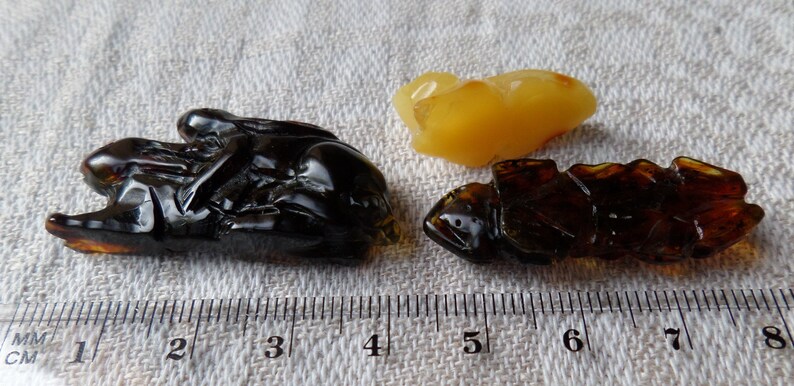 May include: Three amber carvings of insects, one is a light yellow color, and the other two are dark brown. The carvings are displayed on a white fabric surface with a ruler for scale.