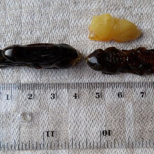 May include: Three amber insect carvings, two are dark brown and one is light yellow. The carvings are approximately 4 cm long and are displayed on a white fabric surface with a ruler for scale.