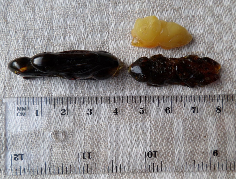 May include: Three amber insect carvings, two are dark brown and one is light yellow. The carvings are approximately 4 cm long and are displayed on a white fabric surface with a ruler for scale.