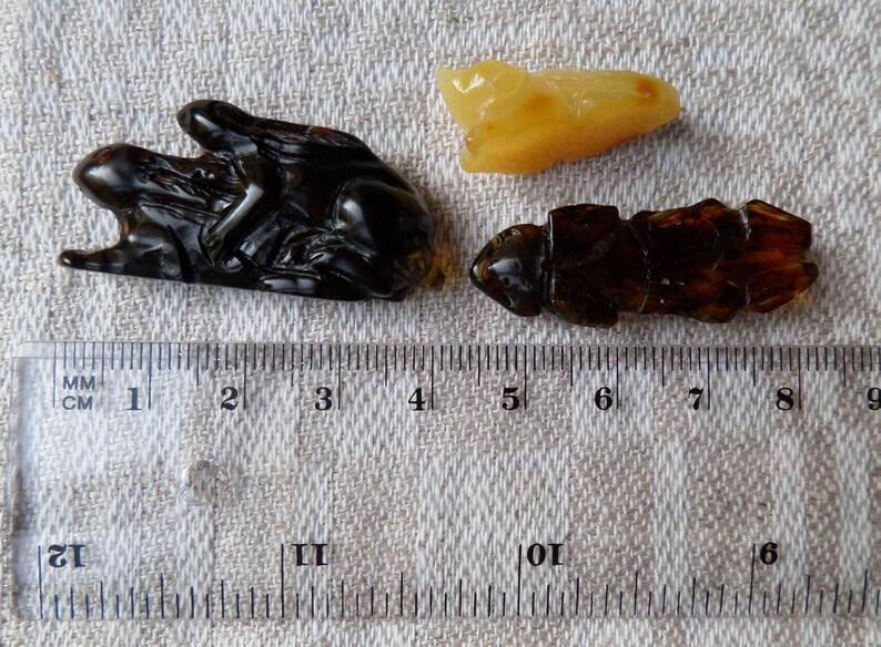 May include: Three amber carvings of insects, one is a dark brown, one is a light brown, and one is a dark brown with a lighter brown insect on top. The carvings are on a white fabric background with a ruler for scale.