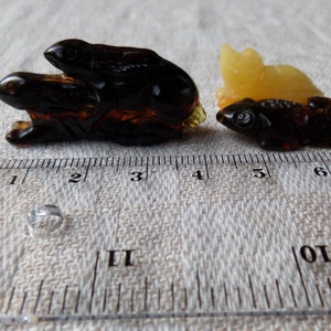 May include: Two amber animal carvings, one of a rabbit and one of a fish, are shown on a white fabric background with a ruler for scale. The rabbit carving is dark brown and the fish carving is light brown.