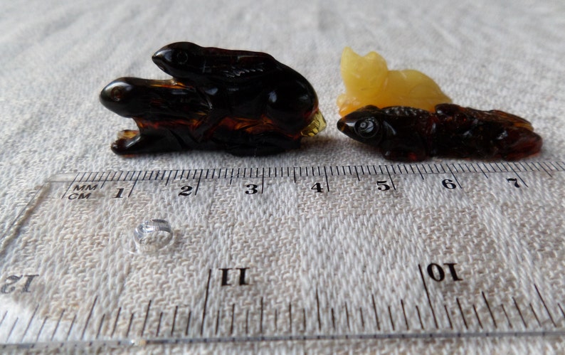 May include: Two amber animal carvings, one of a rabbit and one of a fish, are shown on a white fabric background with a ruler for scale. The rabbit carving is dark brown and the fish carving is light brown.