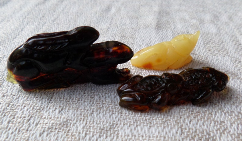 May include: Three amber animal carvings: a dark brown rabbit, a light brown frog, and a light yellow frog. The carvings are detailed and intricate.