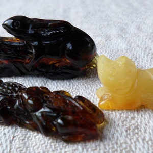 May include: Three amber animal figurines: a dark amber rabbit, a light amber cat, and a dark amber frog. The rabbit and frog are carved in a realistic style, while the cat is more stylized.