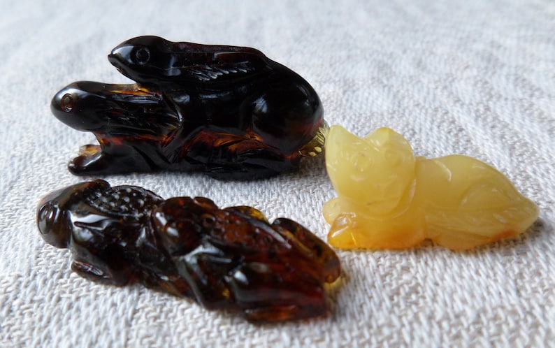 May include: Three amber animal figurines: a dark amber rabbit, a light amber cat, and a dark amber frog. The rabbit and frog are carved in a realistic style, while the cat is more stylized.