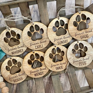 May include: Eight wooden ornaments with a paw print design and the text "forever loved" followed by a dog's name. The names are Whiskey, Carli, Sunday, Shug, Roxii, Wendy, Inkie, and Zeus.