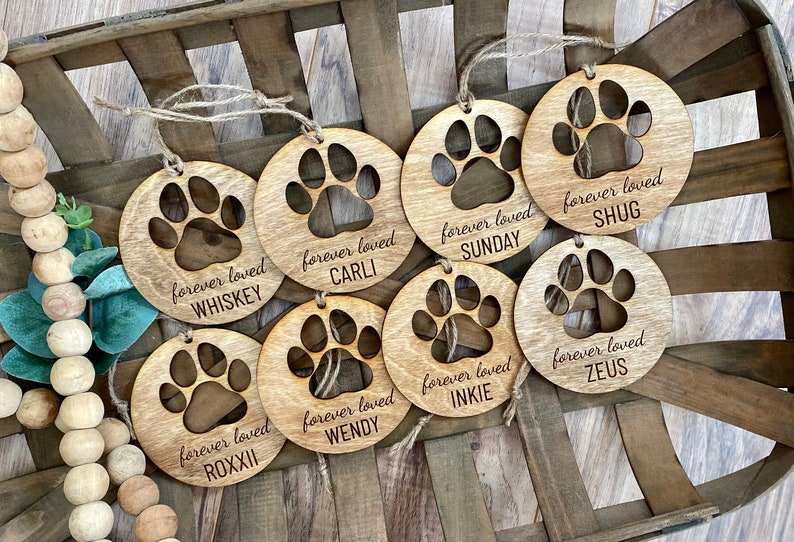 May include: Eight wooden ornaments with a paw print design and the text "forever loved" followed by a dog's name. The names are Whiskey, Carli, Sunday, Shug, Roxii, Wendy, Inkie, and Zeus.