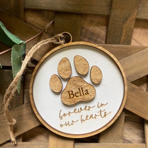 May include: A white wooden ornament with a brown paw print and the name "Bella" engraved on it. The ornament also has the text "forever in our hearts" engraved on it.