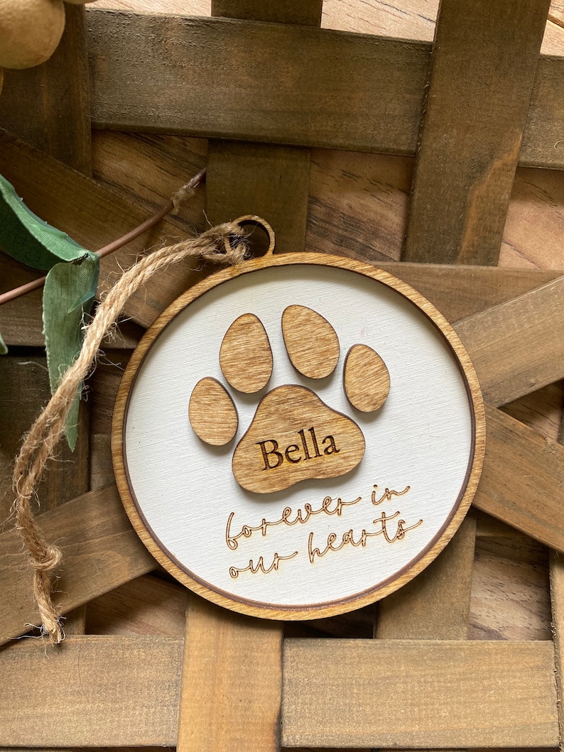 May include: A white wooden ornament with a brown paw print and the name "Bella" engraved on it. The ornament also has the text "forever in our hearts" engraved on it.