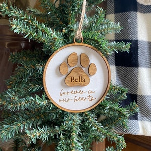 May include: A wooden Christmas ornament with a white background and a brown paw print. The ornament is engraved with the name "Bella" and the text "forever in our hearts".