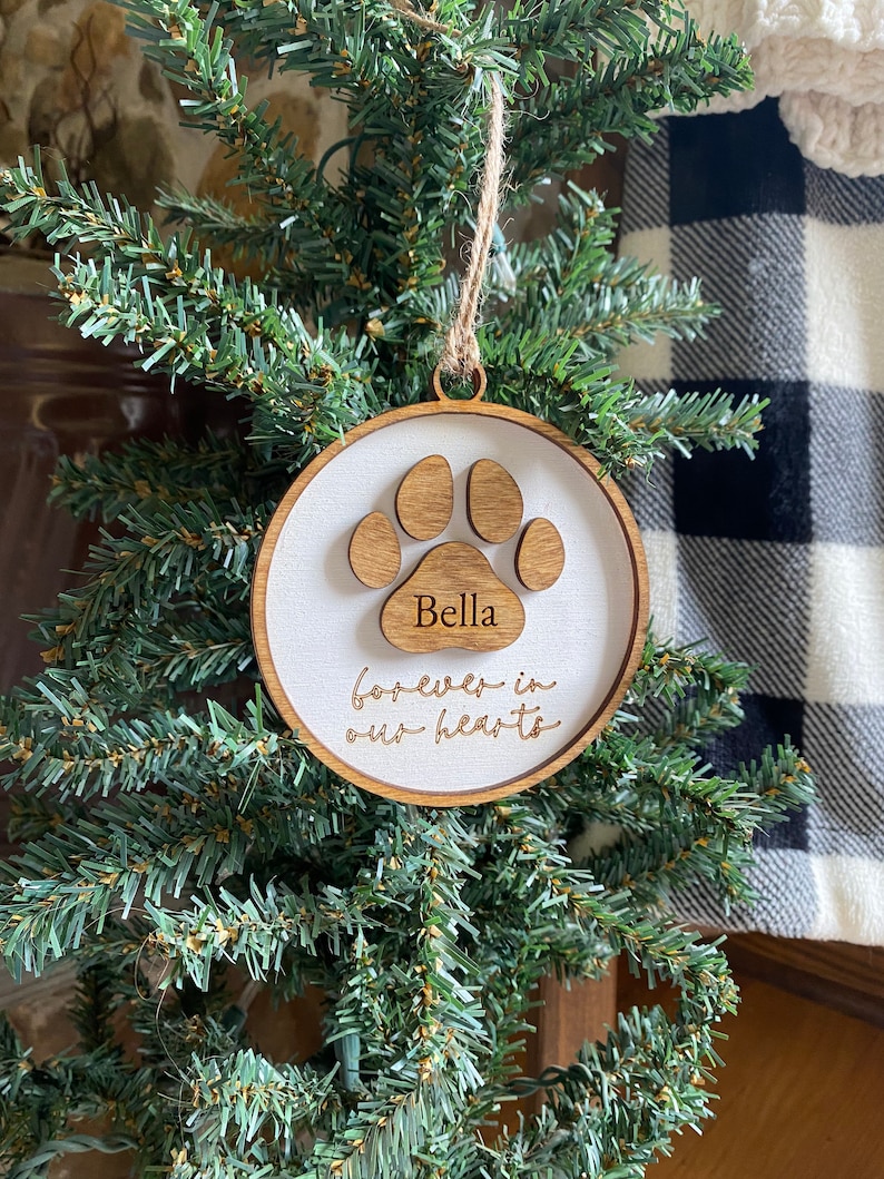 May include: A wooden Christmas ornament with a white background and a brown paw print. The ornament is engraved with the name "Bella" and the text "forever in our hearts".