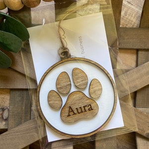 May include: A wooden paw print ornament with the name "Aura" engraved on it. The ornament is white with a brown paw print and is packaged in a clear plastic bag.