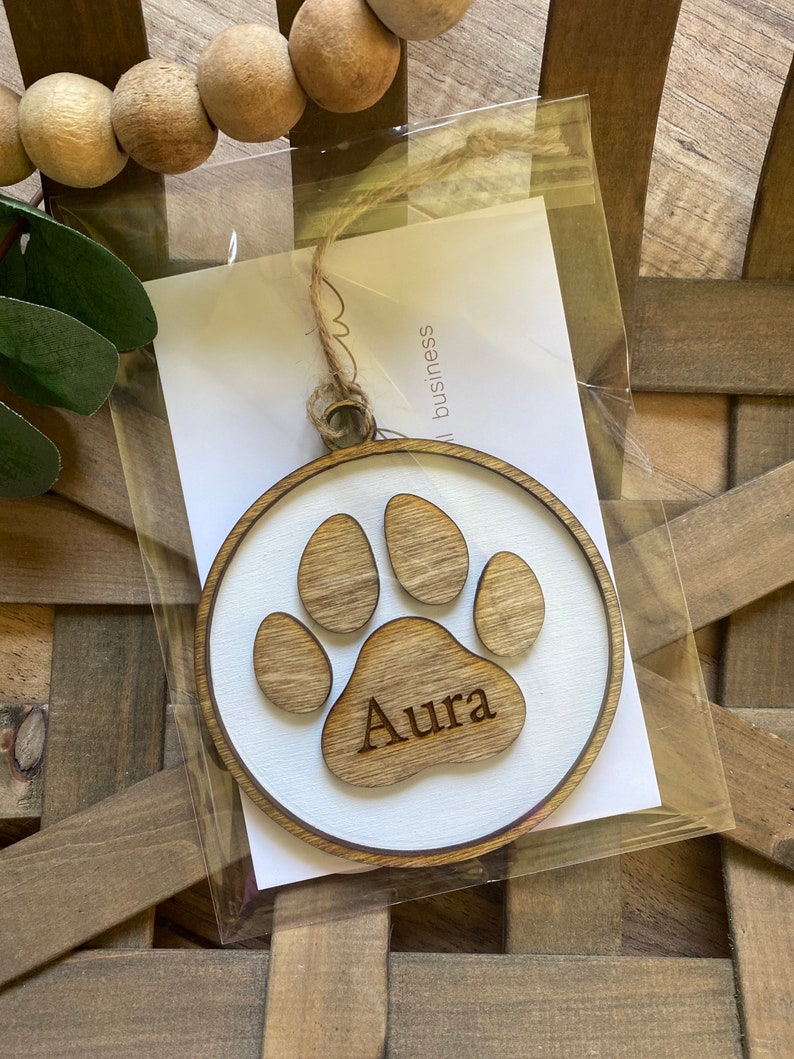 May include: A wooden paw print ornament with the name "Aura" engraved on it. The ornament is white with a brown paw print and is packaged in a clear plastic bag.